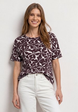 Femme aux longs cheveux portant une chemise à manches courtes à motif floral foncé et un pantalon blanc, souriante devant un mur blanc.