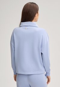 Donna con lunghi capelli castani indossa una maglietta a maniche lunghe blu chiaro e pantaloni coordinati, in piedi di spalle su uno sfondo neutro.