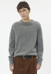 Pulover gri tricotat cu un guler rotund tip țesătură, asortat cu pantaloni din bumbac de culoare maro. Puloverul are mâneci lungi și o croială lejeră.