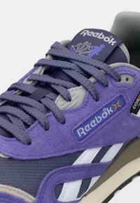 Purpura un pelēka Reebok sneaker ar teksturētām šņorēm, zamšādas un sieta paneļiem, kā arī redzamām zīmola ikonām, tostarp Apvienotās Karalistes karogs.