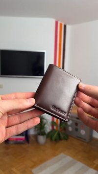 Hnědá kožená peněženka s hladkým povrchem, designem na dvě sklady a embosovaným logem. Držena mezi dvěma rukama na pozadí v interiéru.