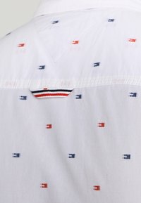 Camisa de tela blanca con un patrón repetido de pequeños cuadrados rojos y marinos. Presenta un acento a rayas cerca del escote posterior.
