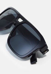 Occhiali da sole neri in plastica con grandi lenti quadrate, dotati di lenti scure e di un design del telaio minimalista. Accentuati da una piccola cerniera.