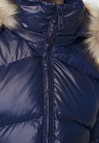 Marinblå puffjacka med dragkedja, hög krage och fuskpälsdetalj. Har kviltade partier och en slät, glänsande tygtextur.