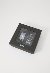 Čierna krabička s priehľadným oknom, vo vnútri sú dva strieborné opaskové spony, jedna obdĺžniková a jedna s kolíkom, s logom "BOSS" pod oknom.