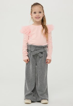 Top lungo a maniche lunghe di un tenue rosa pallido con dettagli a balze sulle spalle, abbinato a pantaloni grigi a gamba larga con vita regolabile. Scarpe semplici completano l'outfit.