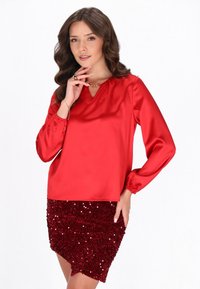 Blusa rossa di satin a maniche lunghe con scollatura a V e polsini elasticizzati, abbinata a una mini gonna rossa glitterata con strass e orlo asimmetrico.