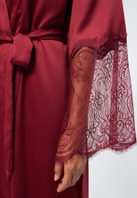 Μπορντό σατέν ρόμπα με δεμένη ζώνη στη μέση και διαφανή μανίκια από δαντέλα με λουλούδια που εμφανίζουν ένα χέρι, πάνω σε λευκό φόντο.