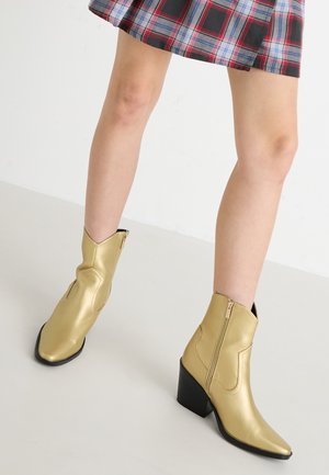 Gouden metallic enkellaarzen met puntige neuzen, ritsluiting en een blokhak. Gladde structuur met gestikte accenten en een klassieke vorm.