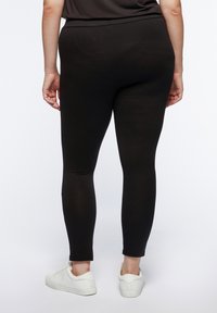 Leggings neri realizzati in materiale elasticizzato, design aderente, lunghezza alla caviglia, abbinati a sneakers bianche. Leggera lucentezza senza motivi visibili.
