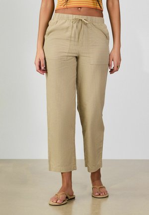 Žena oblečená v béžových krátkych nohaviciach s prednými vreckami a šnúrkou v páse, v kombinácii s béžovými sandálmi na jednoduchom pozadí.