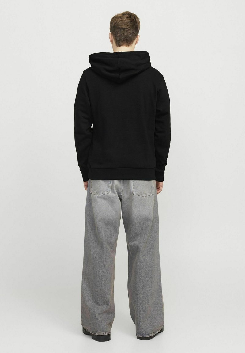 Felpa nera con cappuccio e polsini a coste, abbinata a jeans grigi a gamba larga con tasche posteriori e una texture sbiadita.