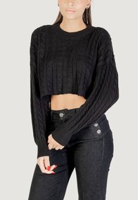 Donna che indossa un maglione corto a treccia nera e pantaloni neri a vita alta, in posa con le mani unite davanti su uno sfondo chiaro.