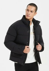 Doudoune noire avec un col montant, des fermetures à bouton et un design matelassé. Portée par-dessus une chemise blanche, présentant une texture lisse et des accents minimalistes.
