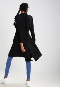 Black long coat with a belted waist and open front, made of smooth fabric; paired with light blue jeans and white sneakers.