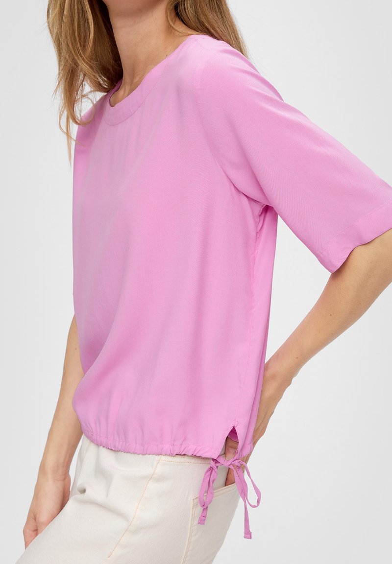 Blouse rose à manches courtes en tissu léger, dotée d'un col arrondi et de liens réglables à l'ourlet pour un ajustement personnalisé.
