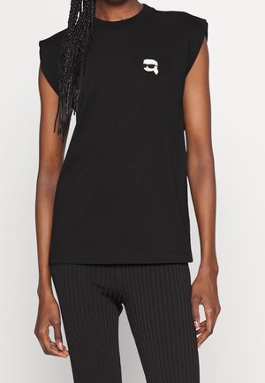 Zwarte mouwloze top met gevoerde schouders, voorzien van een klein wit gezicht met zonnebril als embleem op de linkerborst, gedragen met zwarte geribbelde broek.