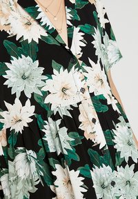 Blommig klänning med svart bakgrund och stora vita och gröna blommor, korta ärmar, knäppning framtill och en samlad midja.