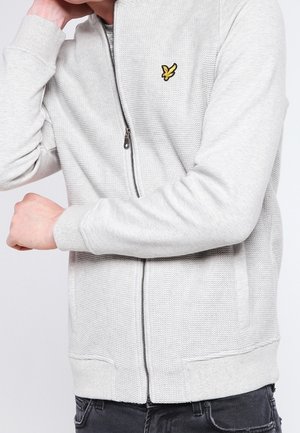 Mężczyzna ubrany w jasnoszarą, zapinaną na zamek, teksturowaną kurtkę z żółtym logo ptaka na piersi, zgiętą ręką dotykającą szyi, widoczne czarne dżinsy.