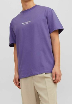 Person trägt ein lila T-Shirt mit der Aufschrift "Originals Studio" und beige Hosen, steht vor einem schlichten hellgrauen Hintergrund.