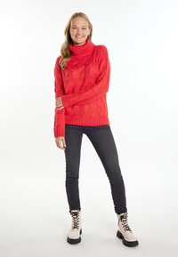 Rode gebreide coltrui met een kabelbreipatroon, lange mouwen en geribbelde zoom, gecombineerd met zwarte skinny jeans en beige veterschoenen.
