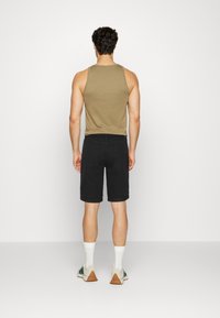 Homme debout de dos, portant un débardeur marron, un short noir, des chaussettes blanches et des baskets blanches avec des semelles marron.