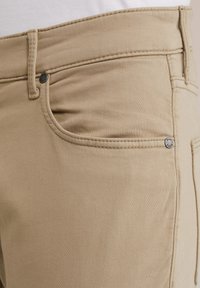 Beiga bomullbyxor med en mjuk textur, med en framficka och en metallknapp som accent. Sömmarna är synliga längs sidorna.