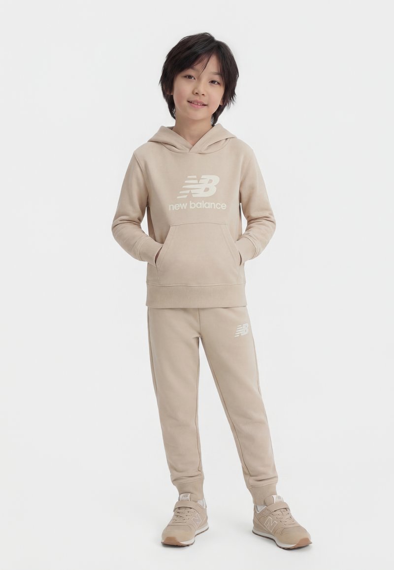 Παιδί που φοράει μπεζ φούτερ με κουκούλα New Balance, ταιριαστό φόρμα και αθλητικά παπούτσια, στέκεται με τα χέρια στις τσέπες του φούτερ μπροστά σε λευκό φόντο.