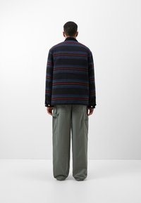 Gestreiftes Baumwollhemd in marineblau, rot und grünen Tönen; entspannter Schnitt mit Kragen und Knopfdetails. Kombiniert mit hellgrünen Cargo-Hosen.