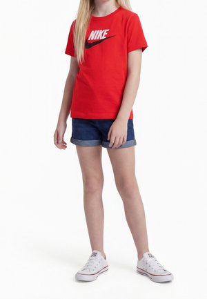 Niño con camiseta roja de Nike, pantalones cortos de mezclilla remangados y zapatillas blancas de pie contra un fondo blanco.