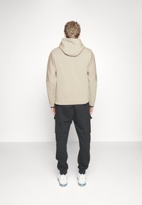 Man som bär en beige huvtröja, svarta cargobyxor och vita sneakers, står vänd mot en vit vägg i en enkel studio-miljö.