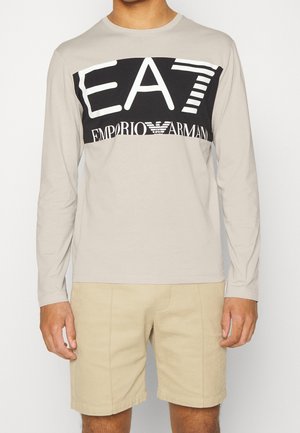 Personne portant une chemise beige à manches longues avec un logo EA7 Emporio Armani noir et blanc sur la poitrine et un short beige clair.