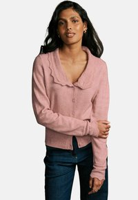 Rožinė megzta džemperis su banginiu apykakle, mygtukų užtrauktuku ir raukšlėmis ant rankovių; dėvima ant tamsių džinsų. Matoma minkšta tekstūra.