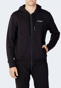 Haut à capuche noir zippé en mélange de coton, avec une capuche, des poches kangourou et un logo sur la poitrine. Texture lisse et coupe régulière.