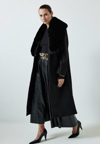 Vrouw in een lange zwarte jas met een bontkraag, zwarte geplooide rok, een riem met luipaardprint, zwarte hoge hakken en gouden sieraden op een witte achtergrond.