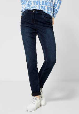 Personne portant un jean bleu foncé avec des revers et des baskets blanches, debout jambes croisées devant un fond uni.
