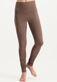Leggings marron taille haute avec une texture lisse, des jambes fuselées et des détails froncés aux chevilles. Présente un petit logo près de la taille.