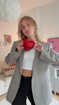 Young woman in a checked blazer and white crop top holds a red sequined heart near her chest in a cozy living room.
