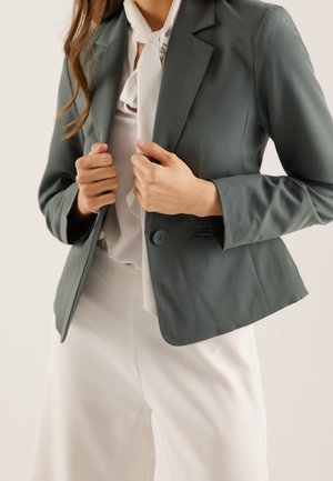 Person, der justerer en mørkegrå blazer over en hvid bluse med sløjfe, kombineret med hvide bukser med høj talje mod en neutral baggrund.