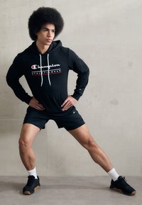 Svart hettegenser med hvite snorer og en rød Champion-logo, kombinert med svarte shorts og svarte sneakers, satt mot en nøytral bakgrunn.