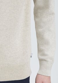 Lys beige genser laget av myk, strukturert strikket stoff. Har ribbestrikkede mansjetter og hem, med en liten etikett synlig på siden.