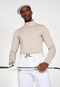 Beeži pika lõhik, millel on veerand tõmblukk, valged aktsendid ja logo. Valge vöö ja golfikinnas, hoides golfikeppi; voolujooneline, sportlik lõike.