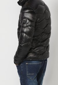 Persona che indossa una giacca imbottita trapuntata nera con una toppa Koroshi sul braccio e jeans di denim blu, con le mani nelle tasche, di profilo.