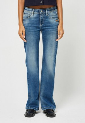 Vrouw draagt vervaagde blauwe bootcut jeans, zwarte schoenen en een korte zwarte top, staand tegen een effen lichte achtergrond.