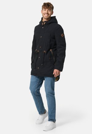 Svart hettejakke med brune detaljer og knapper, med to frontlommer og foret innside, kombinert med blå jeans og hvite joggesko.