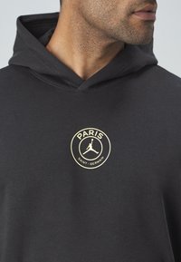 Homme portant un sweat à capuche noir avec le logo du Paris Saint-Germain et la silhouette Jumpman sur la poitrine, face à droite.