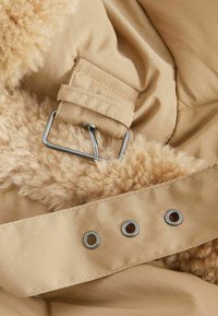 Tissu extérieur beige avec une doublure en sherpa douce de couleur fauve, avec une boucle métallique et trois œillets en métal le long du bord.
