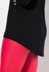 Maglietta nera a maniche lunghe con orlo curvo, abbinata a leggings rosa lucidi e aderenti, con texture liscia e senza motivi visibili.