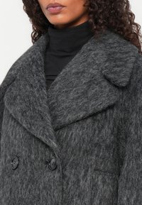 Cappotto grigio in lana con una finitura testurizzata, colletto a scollo ampio, bottoni neri doppi e una tasca singola sul davanti.