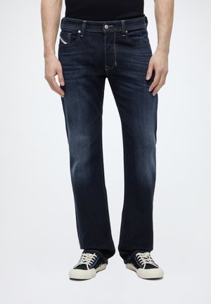 Homme portant un jean droit bleu foncé et des baskets noires et blanches, debout devant un fond clair uni.